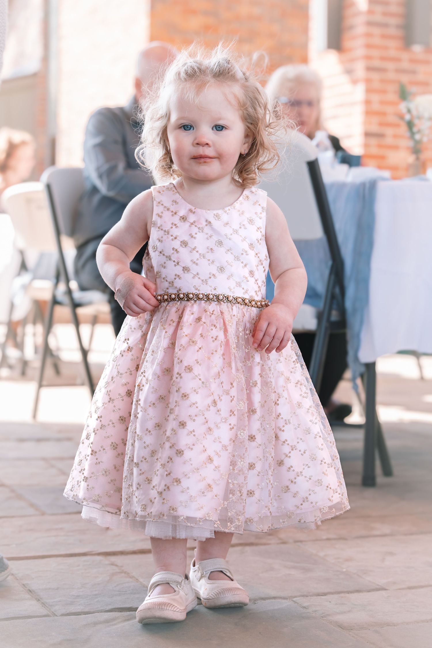 Adorable toddler flower girl in pink lace dress with gold belt at outdoor wedding reception.