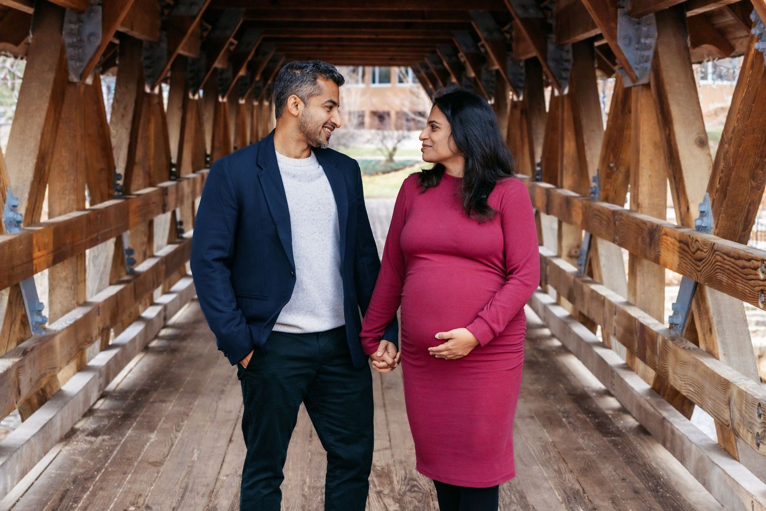 Expecting couple holding hands and smiling at each other inside a rustic wooden covered bridge.