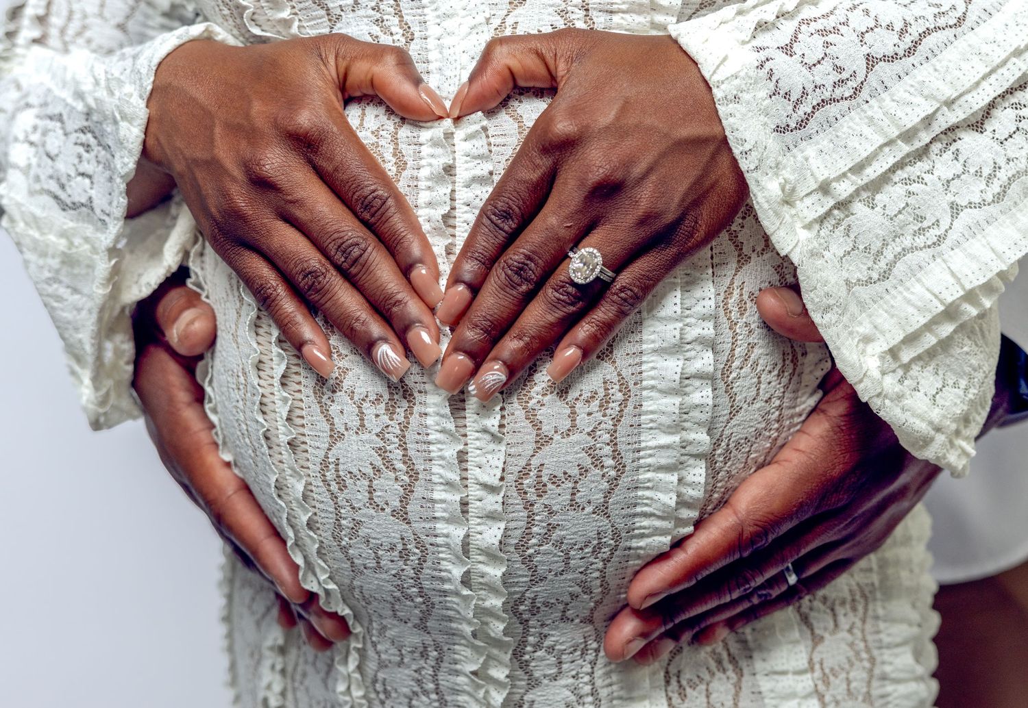 Close-up of couple forming heart shape with hands on pregnant woman's belly in white lace dress.