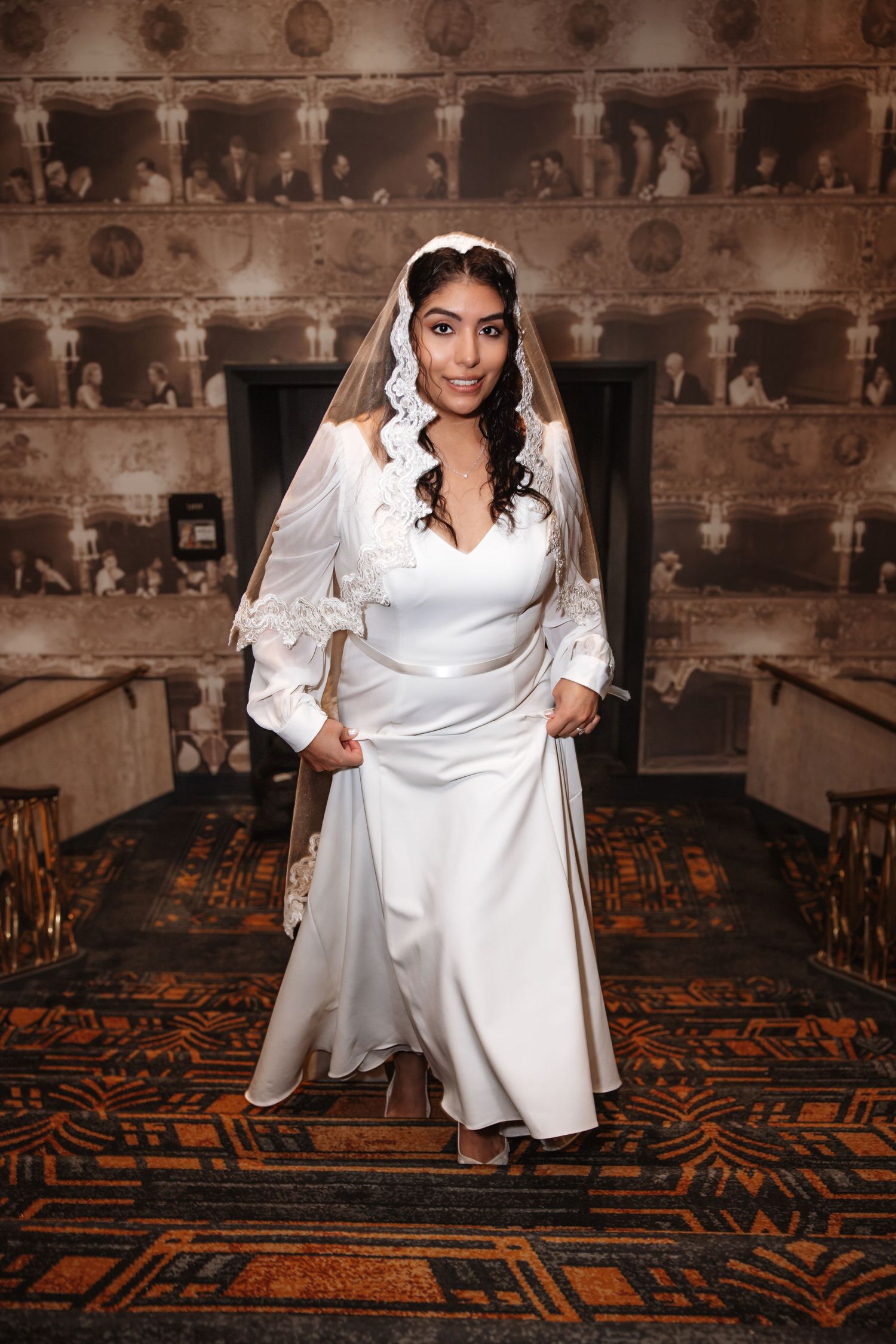 Bride in white satin gown with lace veil poses elegantly in ornate venue with vintage photo wall backdrop.