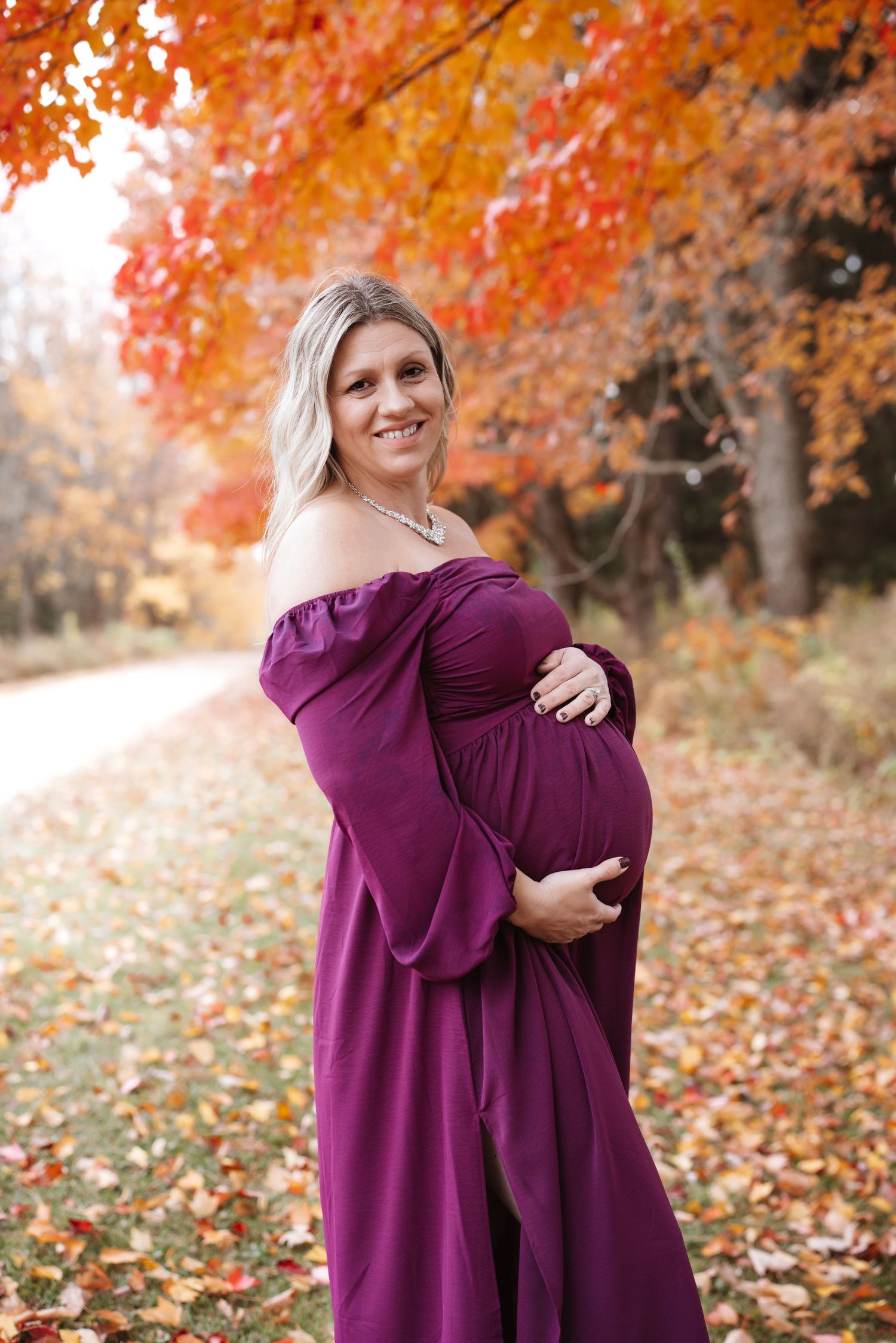 Glowing pregnant woman in purple maternity dress smiling outdoors surrounded by vibrant autumn leaves.
