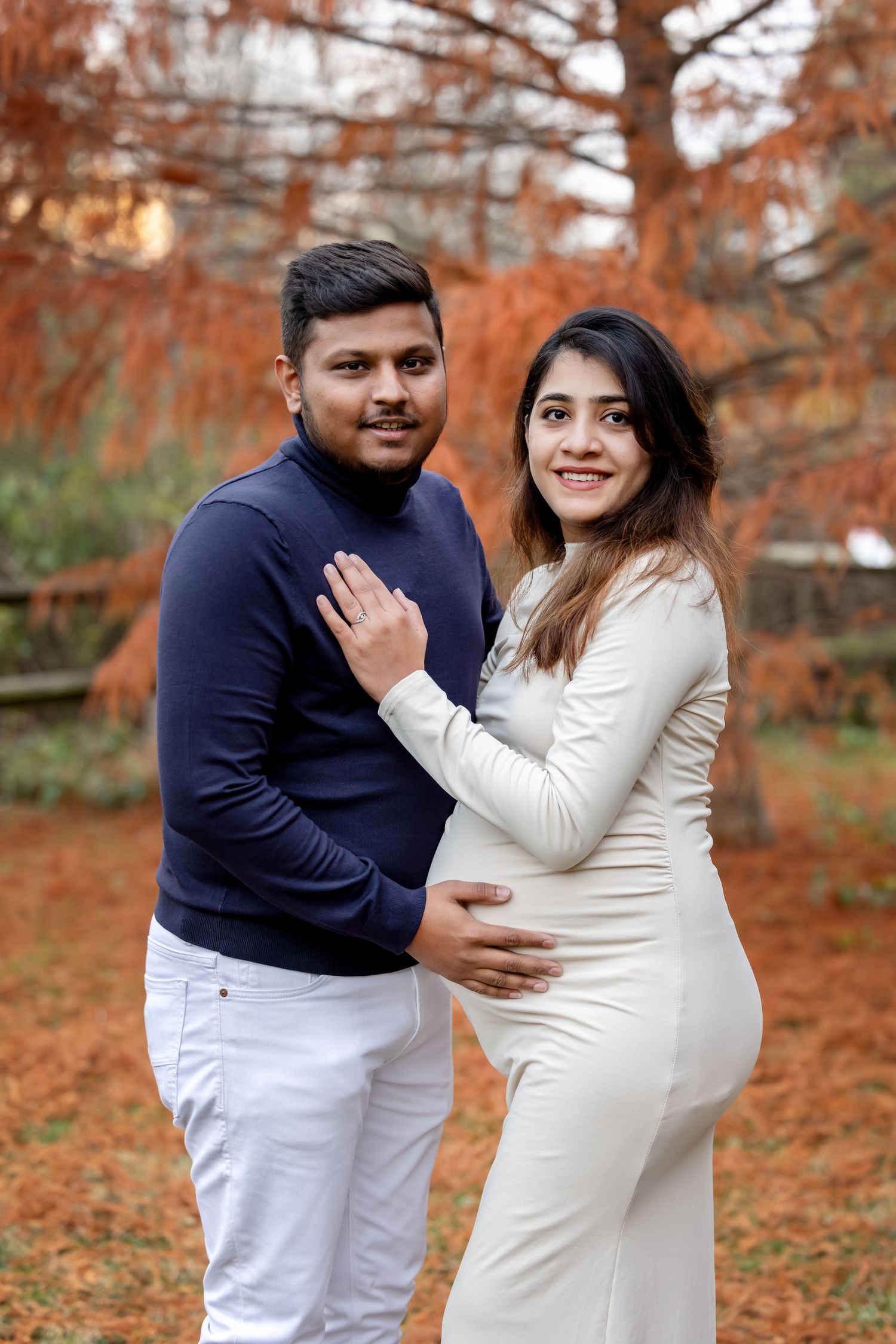 Smiling couple posing outdoors in autumn, pregnant woman in white dress with partner in navy sweater.