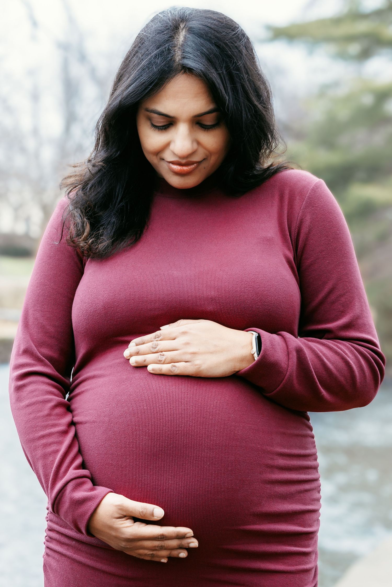 Pregnant woman in burgundy dress lovingly cradling her baby bump with both hands outdoors.