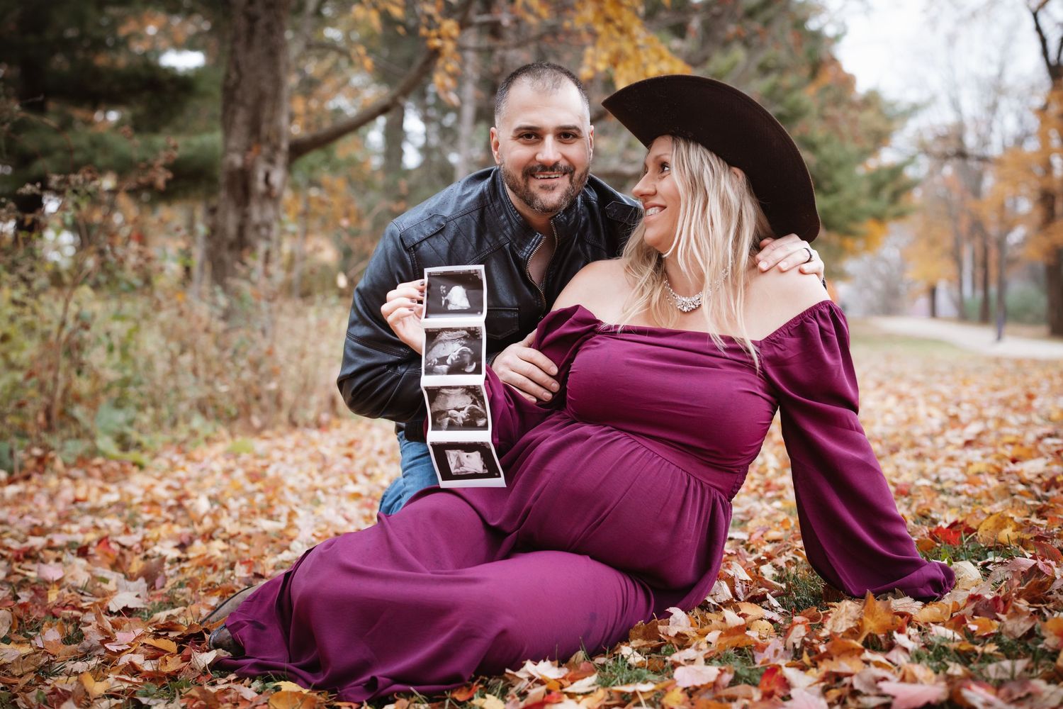 Couple sitting in autumn leaves, pregnant woman in purple dress holding ultrasound photos with smiling partner.