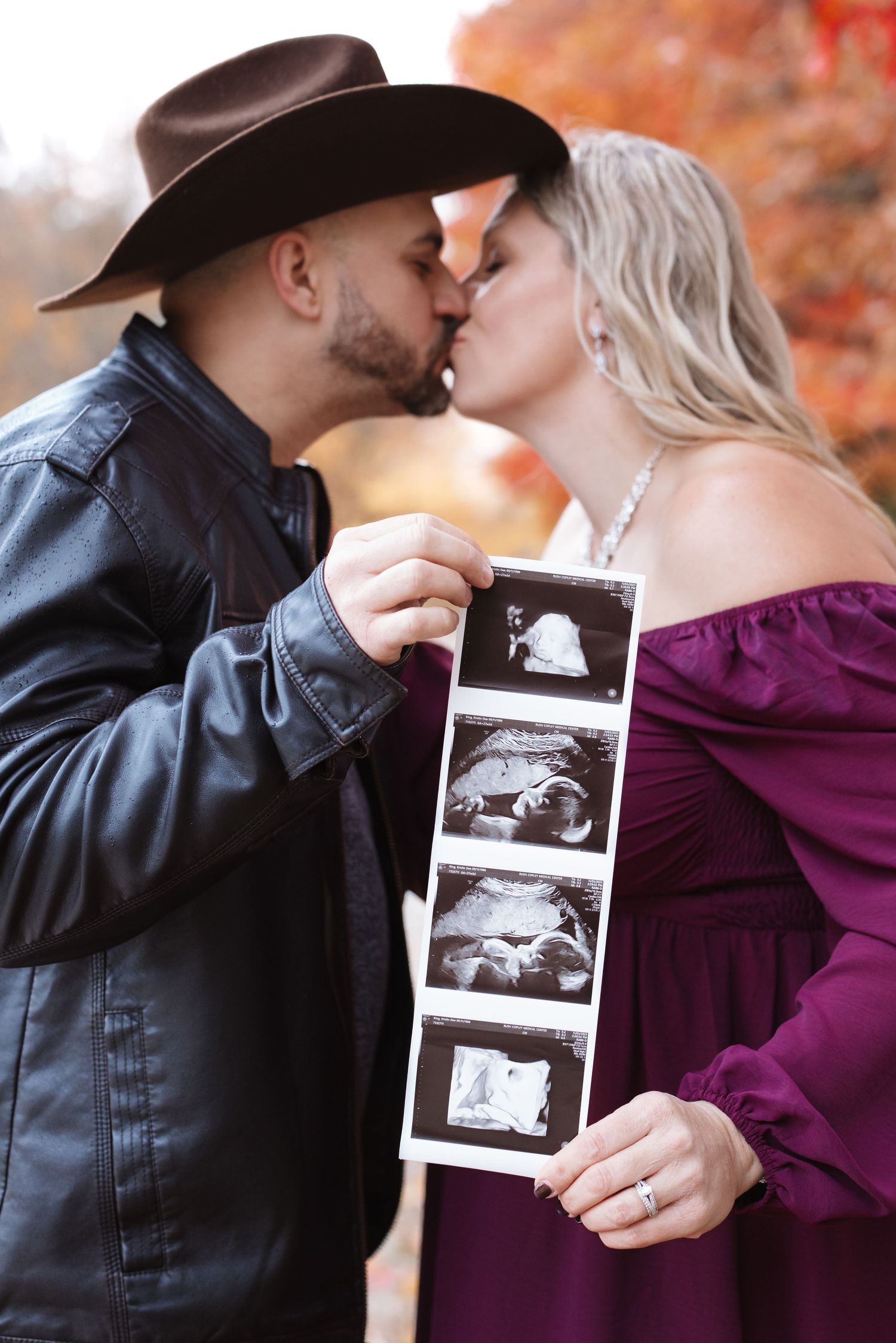 Couple kisses and holds ultrasound photos together against a backdrop of red autumn leaves.