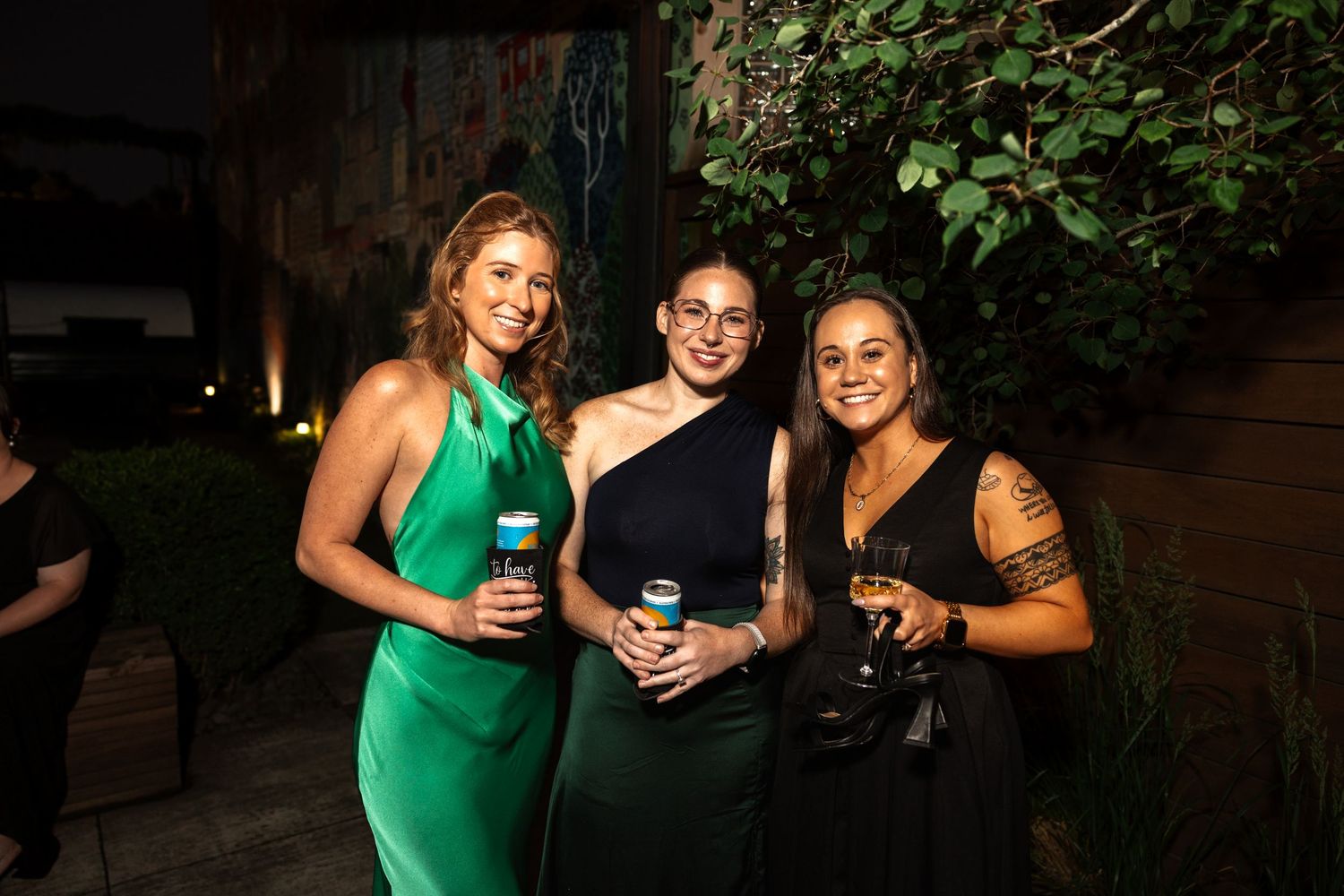 Three smiling women in formal attire holding drinks at an outdoor evening wedding celebration.
