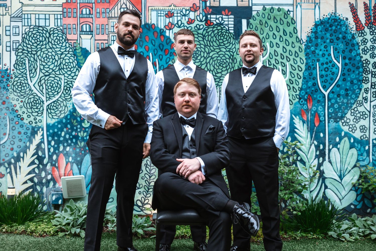 Groom seated with three groomsmen standing behind him in front of vibrant mural.
