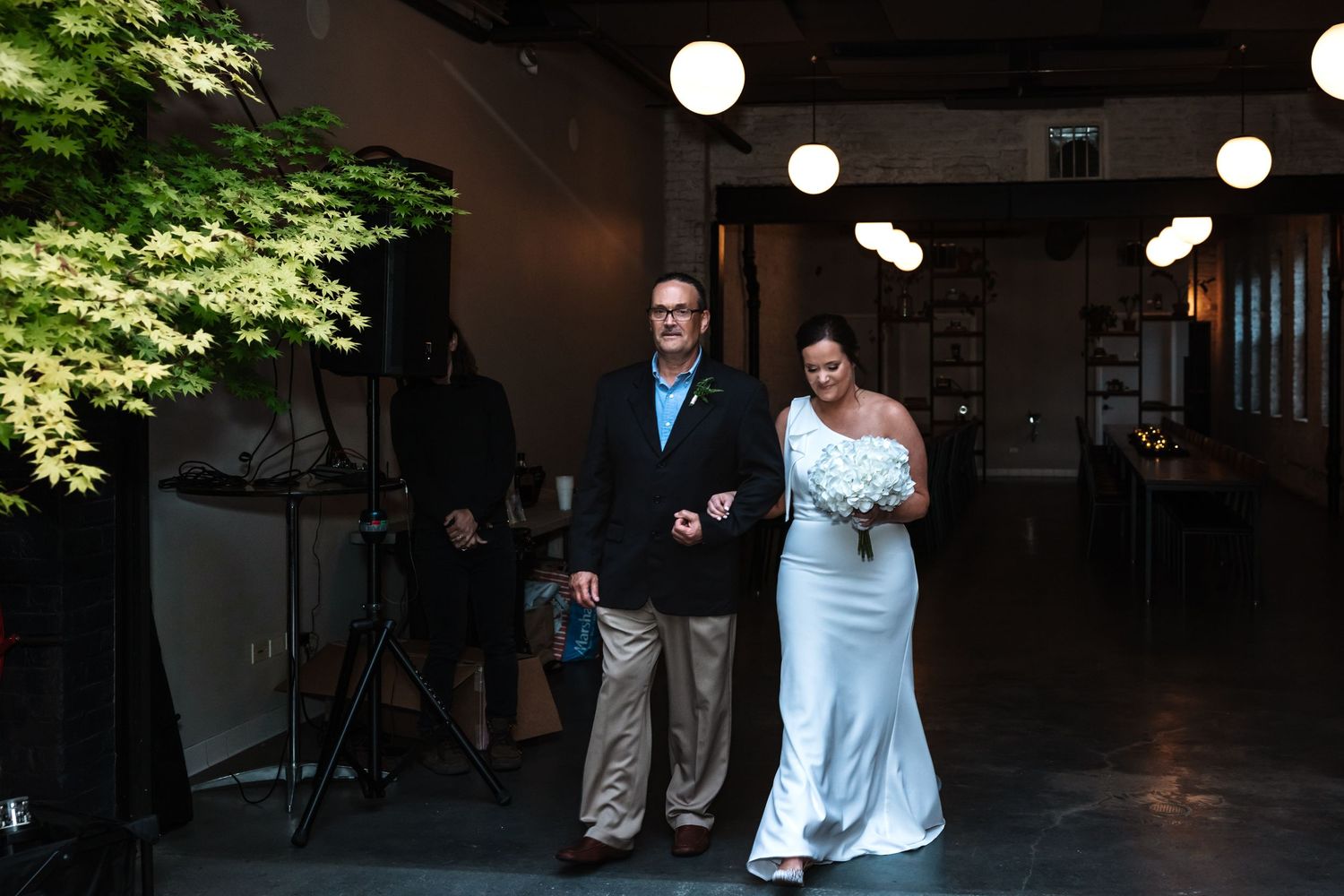 A bride carrying white flowers walks down the aisle with her escort in a modern industrial venue.