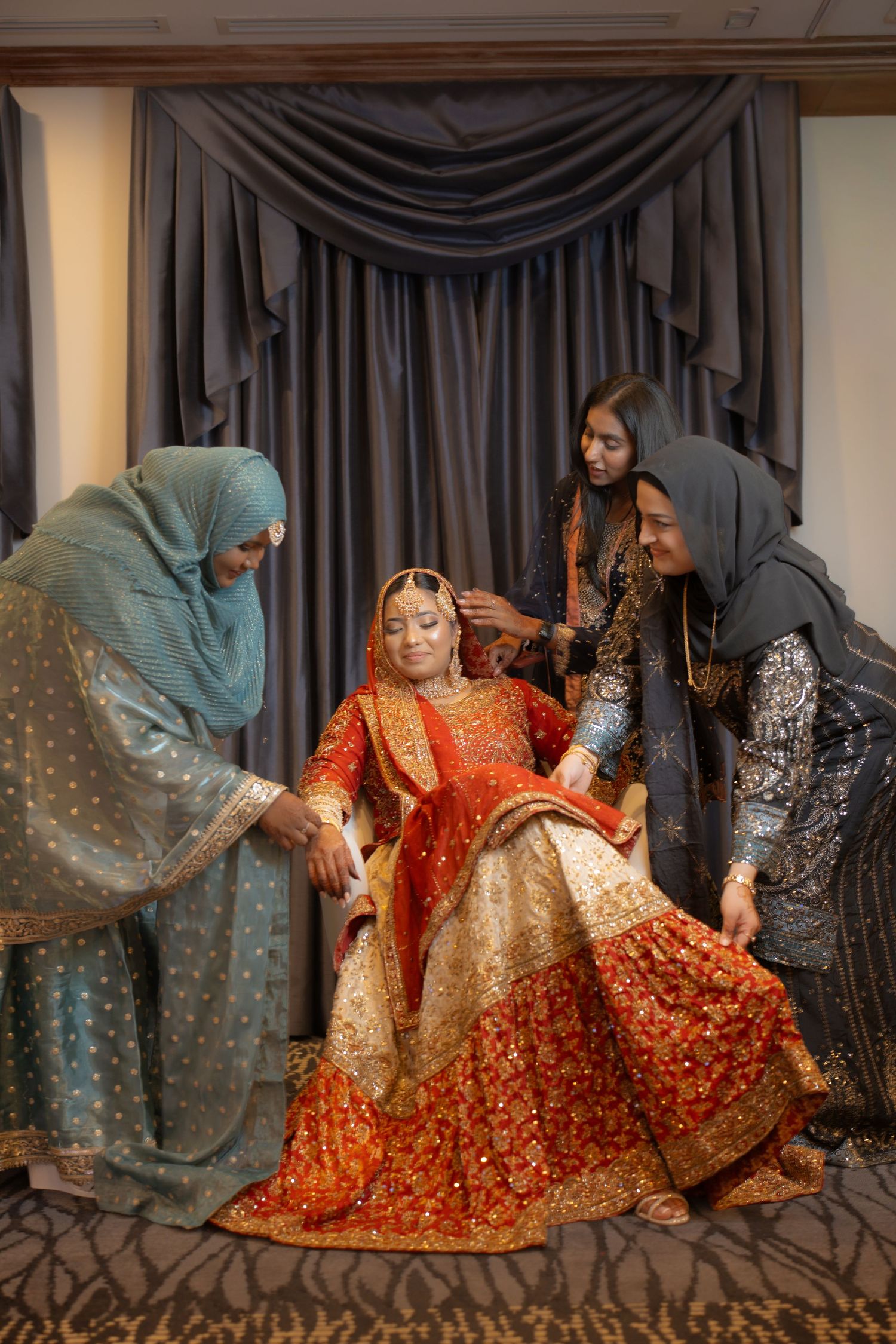 Bride in ornate red and gold lehenga being assisted by three women in hijabs before wedding ceremony.