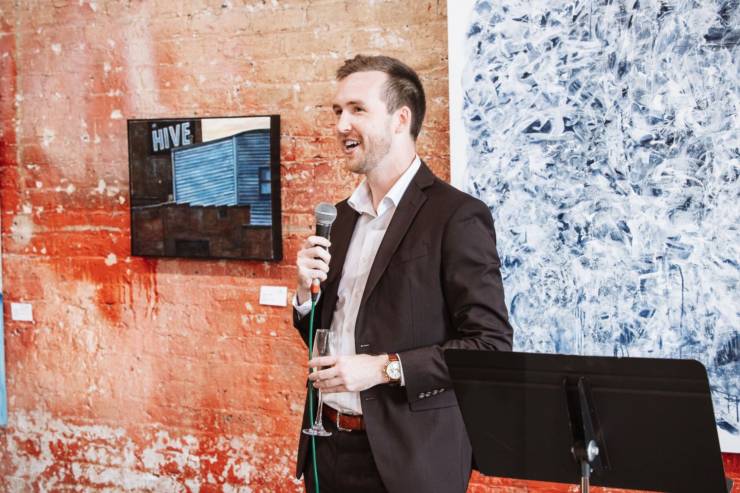 Man in blazer speaking into microphone at Hive event beside abstract artwork on brick wall.