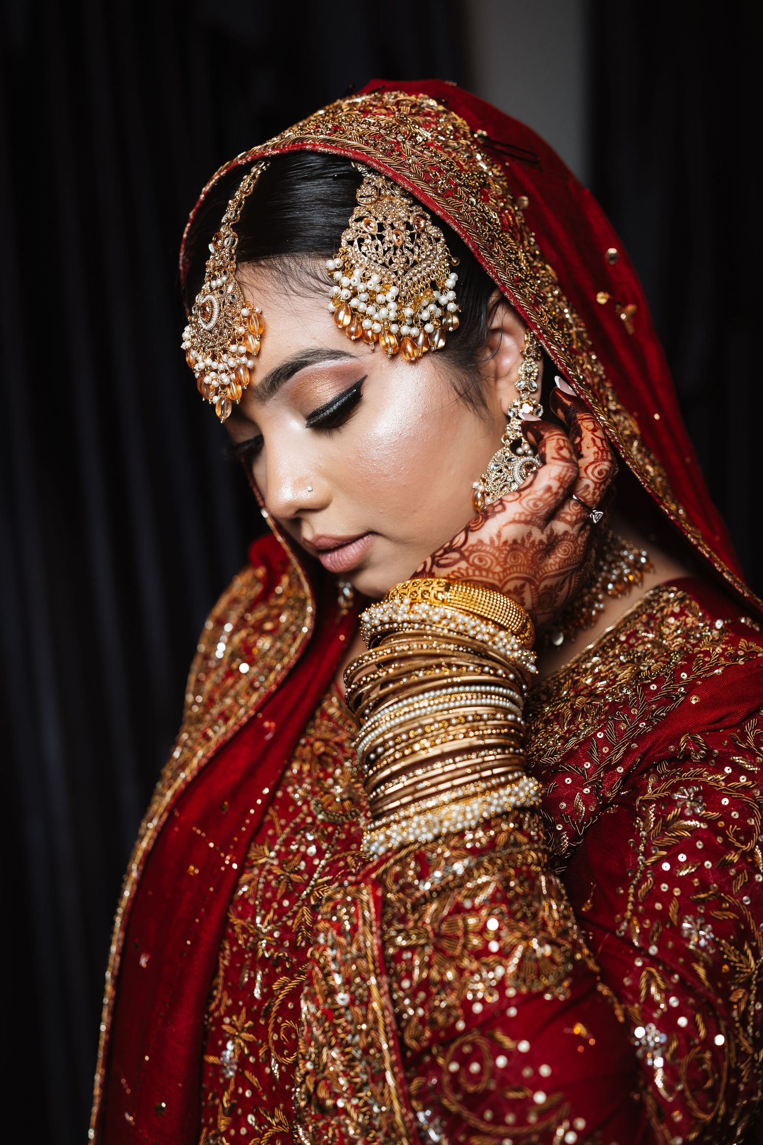 Indian bride in red bridal lehenga with gold maang tikka, bangles, and henna looking down gracefully.