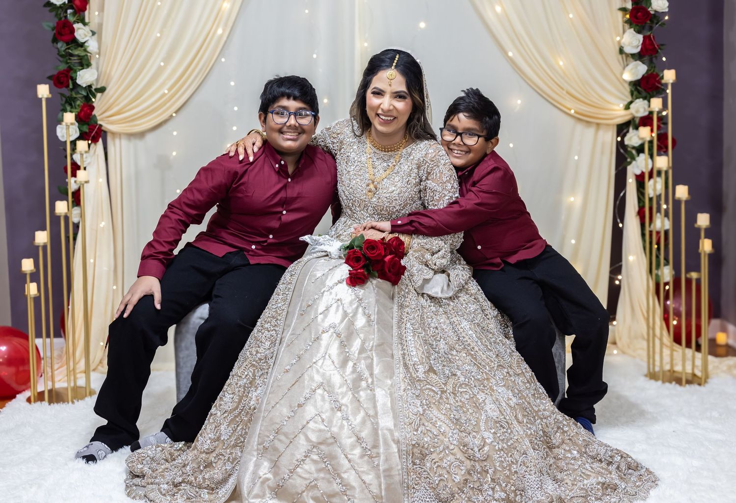 Bride in gold lehenga holds red roses smiling with two young boys in maroon shirts at wedding backdrop.