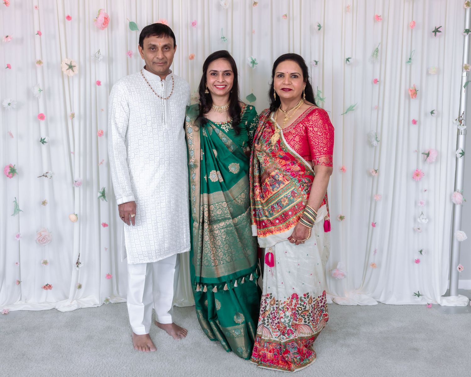 Three family members in vibrant traditional Indian attire pose together at festive celebration with floral backdrop.