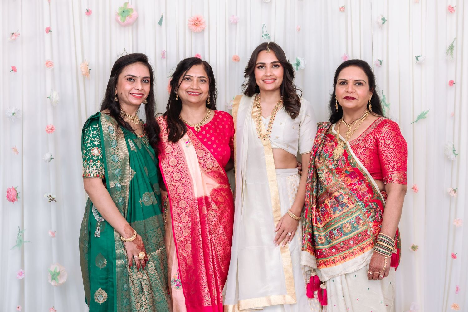 Four women in colorful Indian traditional sarees and lehengas pose together at a decorated wedding celebration backdrop.