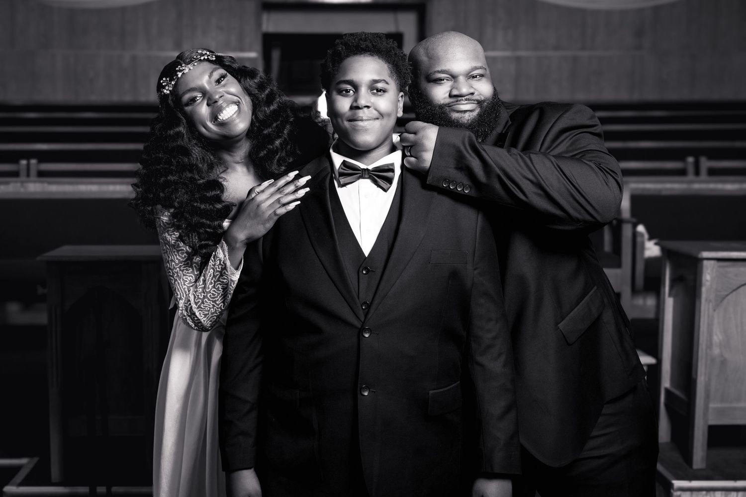 Black and white photo of a family of three posing in a church, young boy in tuxedo flanked by parents.