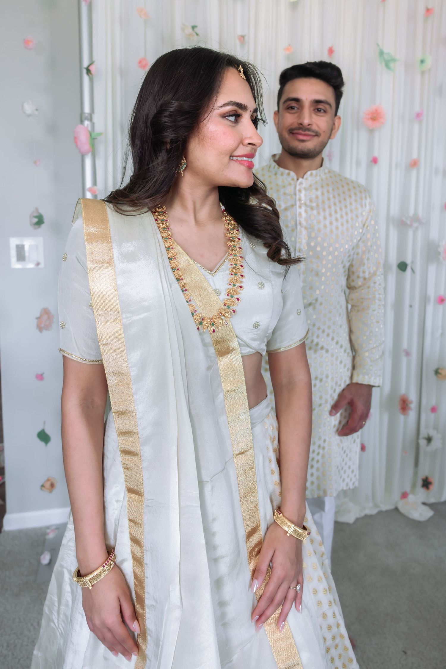 South Asian bride in white lehenga with gold jewelry smiling as groom stands behind her.