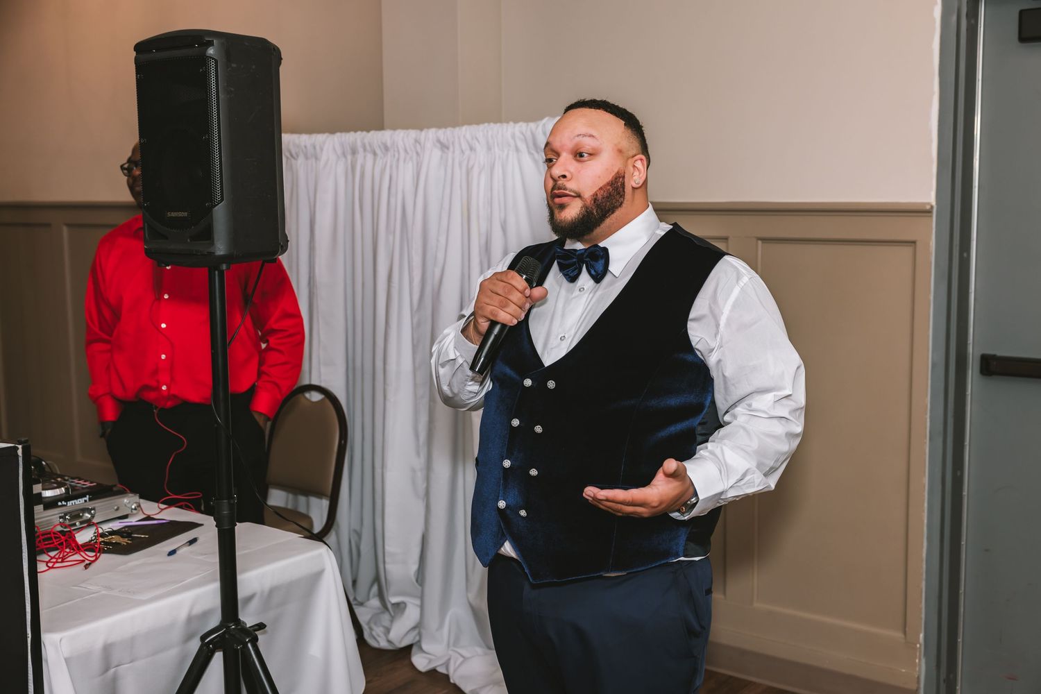 A man in a navy vest and bow tie speaks into a microphone at a wedding reception near a DJ setup.