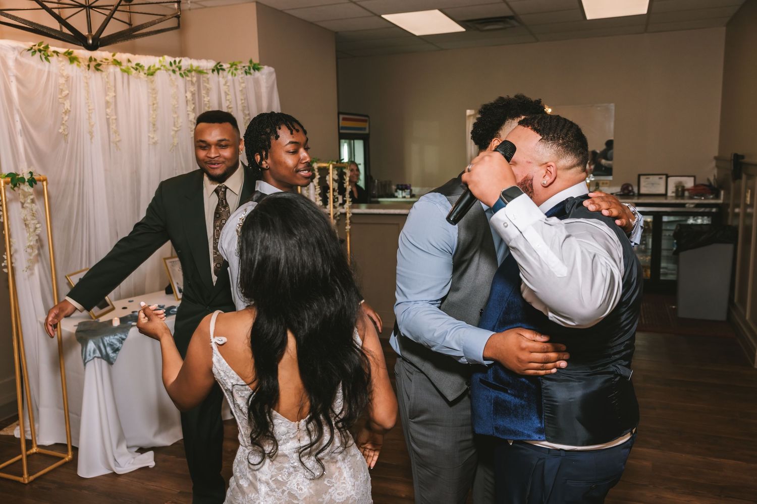 Groom emotionally embracing guest while bride and others celebrate on wedding dance floor.
