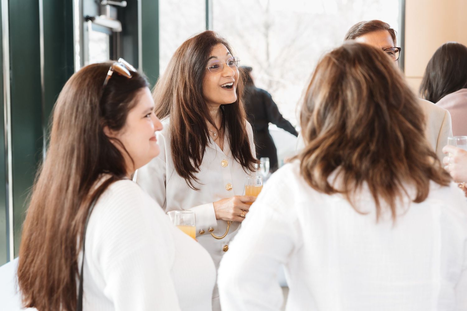 Three women in white attire laugh and socialize while holding drinks at an elegant baby shower.