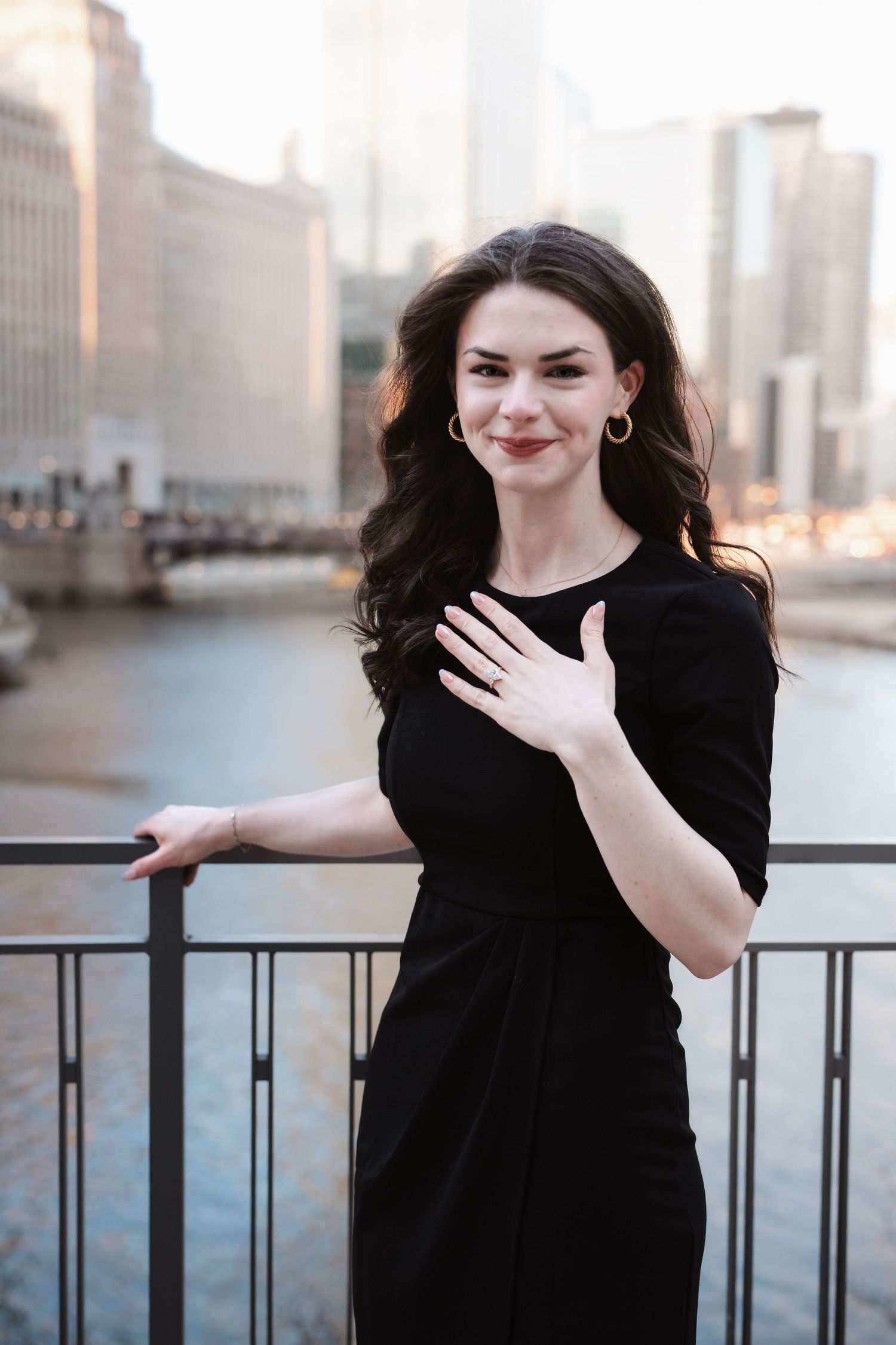 Elegant brunette woman in black dress posing on Chicago Riverwalk bridge with city skyline at dusk.