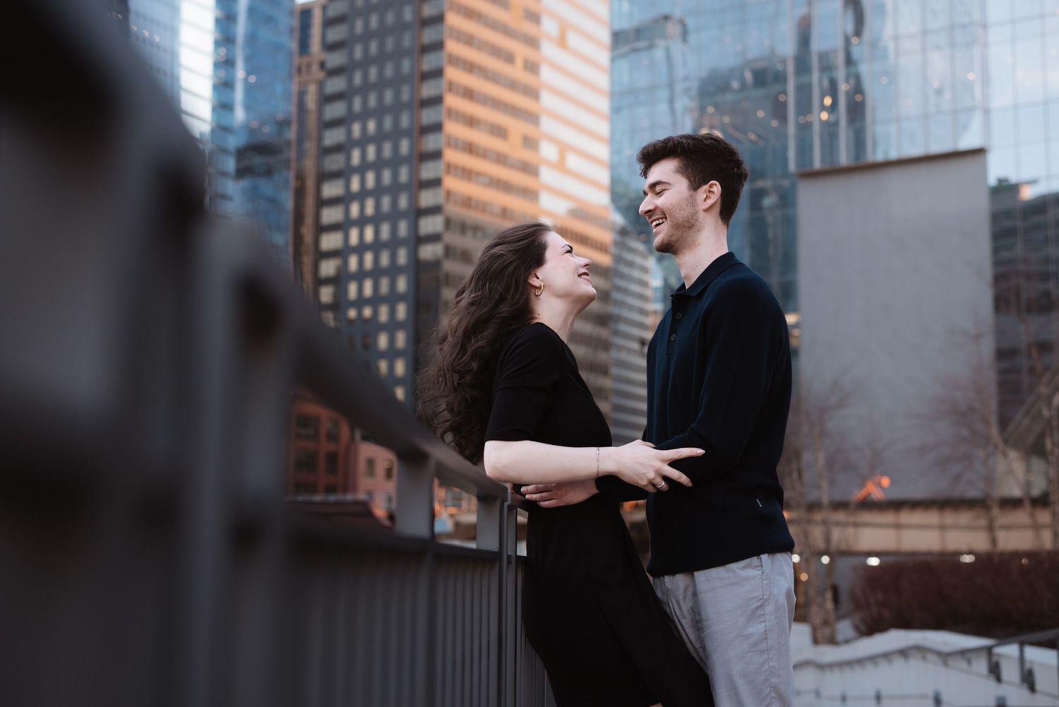 A laughing couple faces each other on a city bridge with tall skyscrapers in the background.