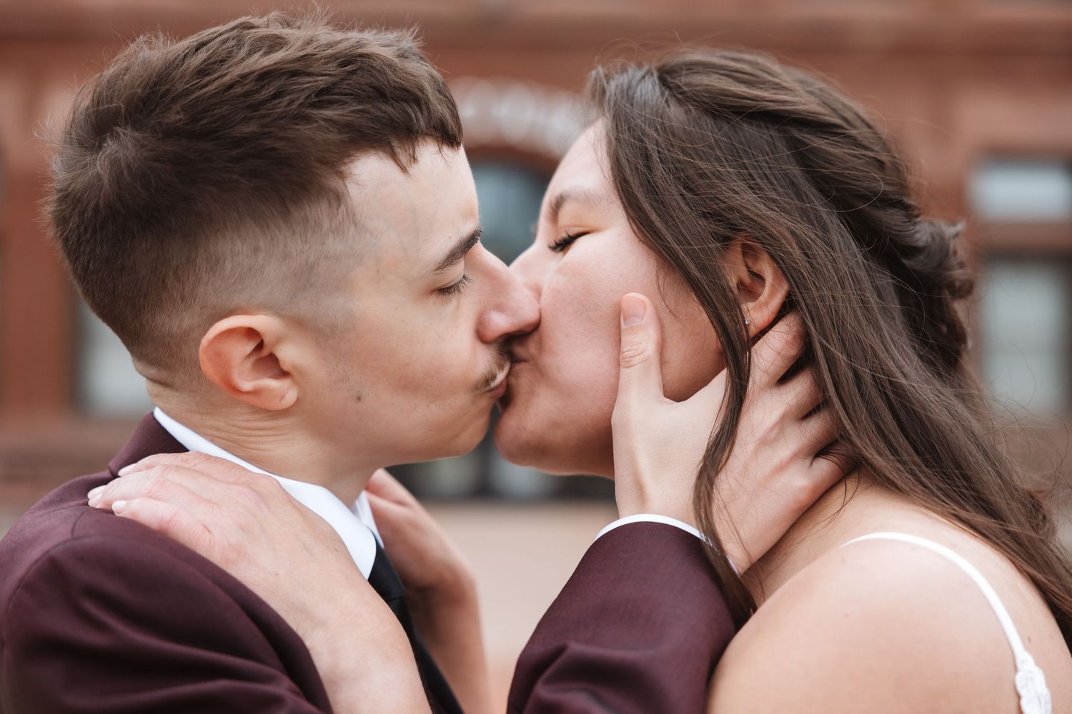 A couple shares a romantic kiss outdoors, with the groom in a burgundy suit and bride in a white dress.