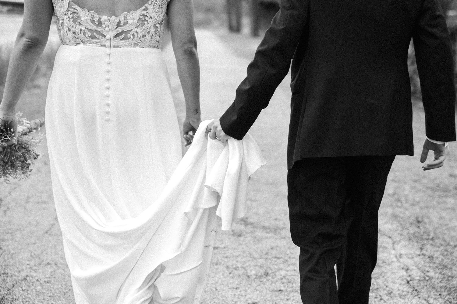Black and white photo of a bride and groom walking hand in hand, groom holding the bride's flowing wedding dress.