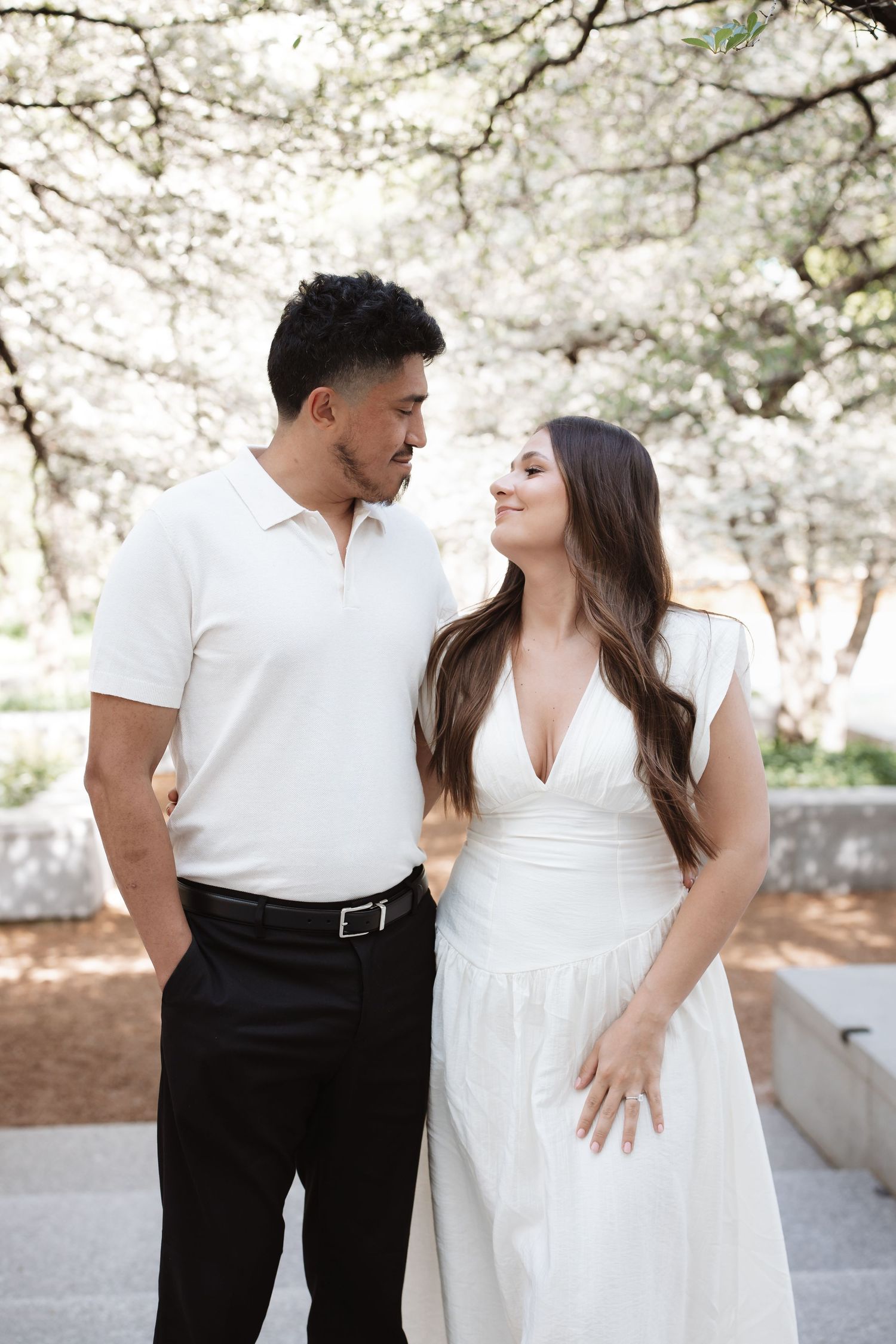 Couple in matching white outfits share a romantic moment under blooming trees in an outdoor park setting.