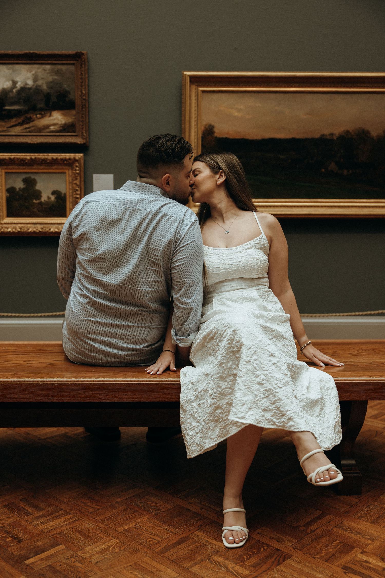 Couple sharing a kiss while seated on a wooden bench surrounded by landscape paintings in a museum.