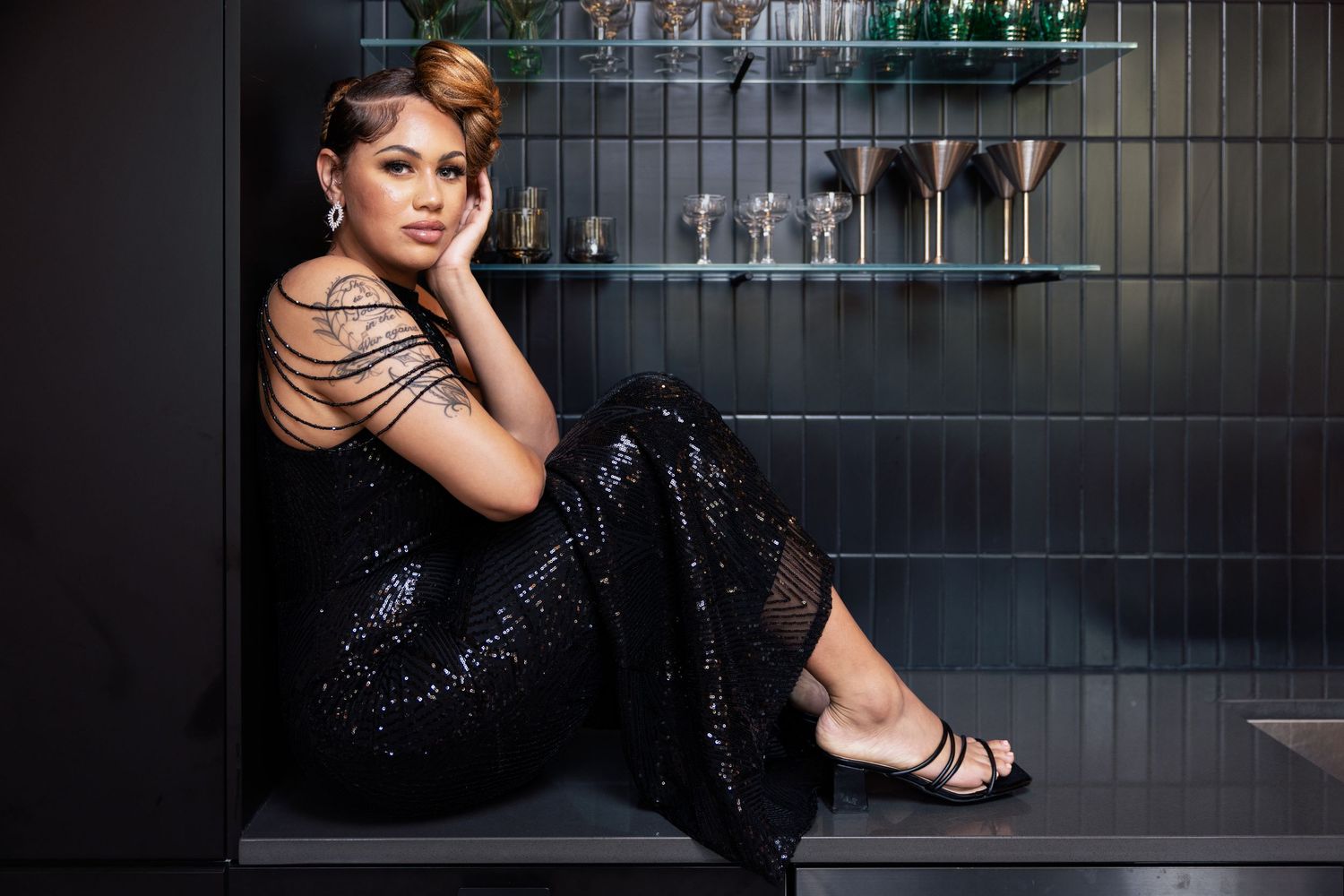 Woman with tattoo in black sequin jumpsuit sitting on floor against dark tiled wall with heels.