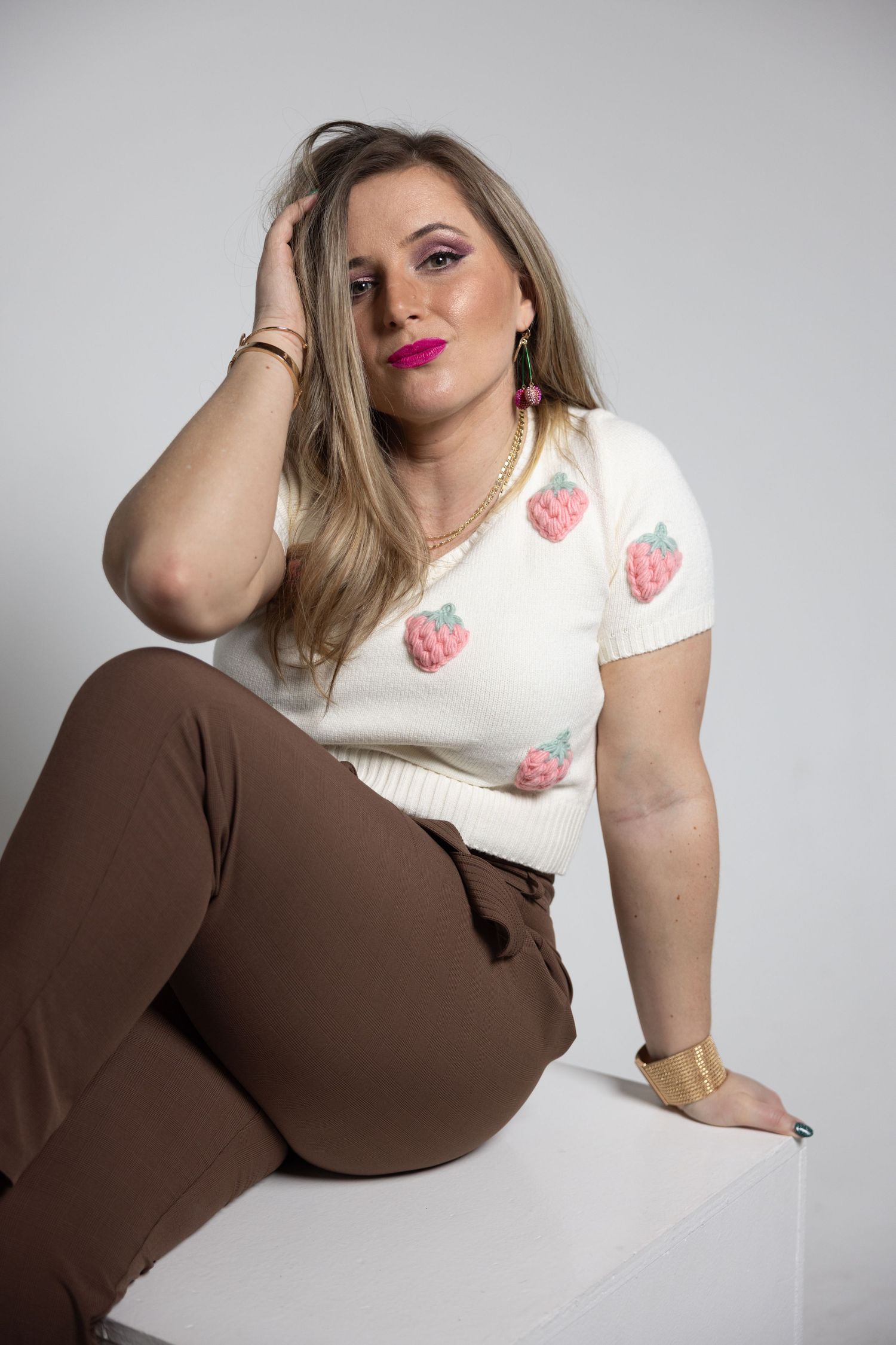 Blonde woman sitting on white cube wearing strawberry print crop top and brown pants with gold accessories.