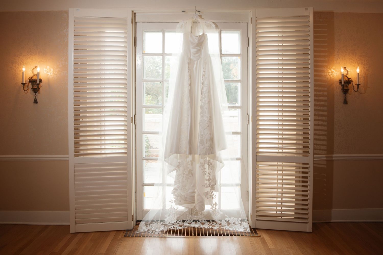 Elegant lace wedding gown hanging between white shuttered French doors with warm wall sconces on each side.
