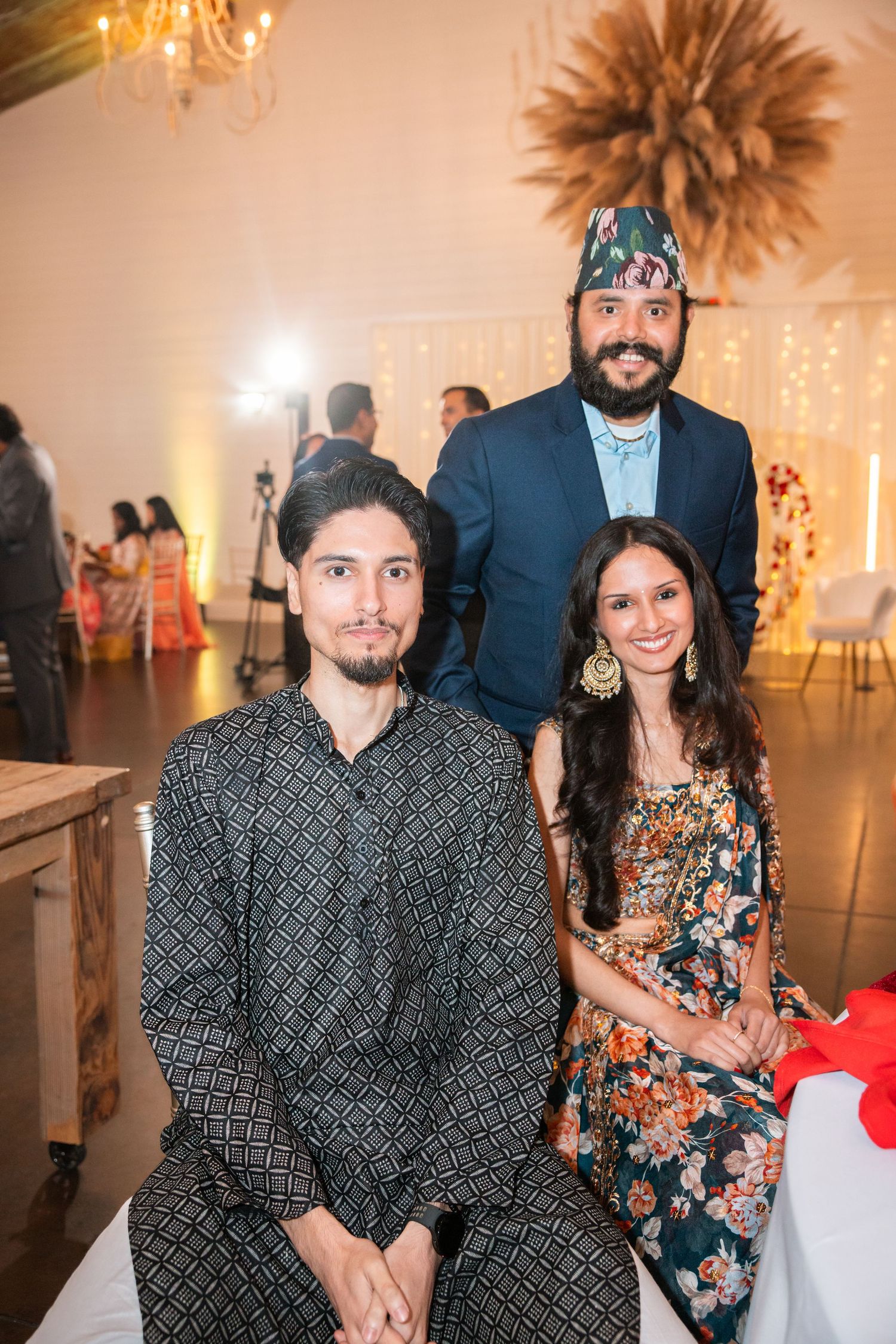 Three guests at a South Asian wedding event, man standing in traditional Nepali hat with two seated friends.