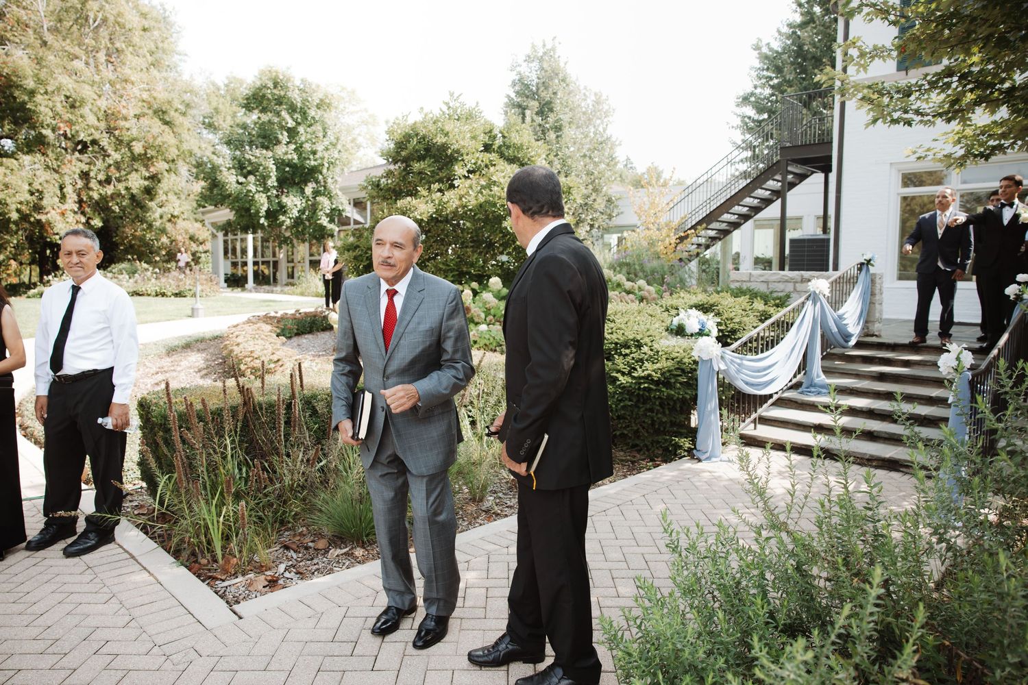 Men in formal suits converse outdoors near a decorated wedding venue entrance with lush greenery.