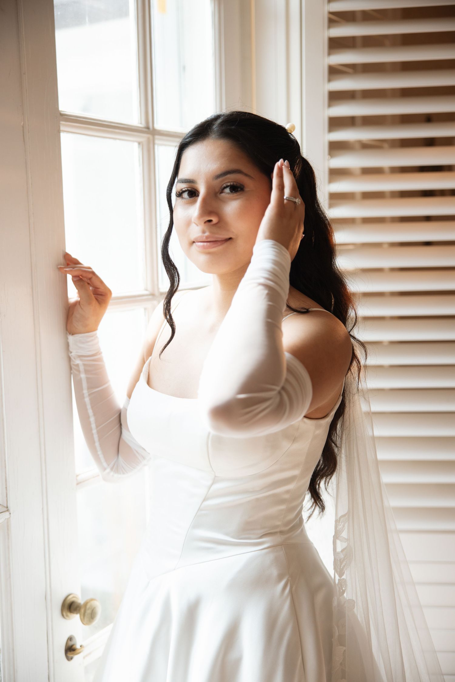 Bride in white wedding dress with sheer gloves poses by window with wooden shutters.