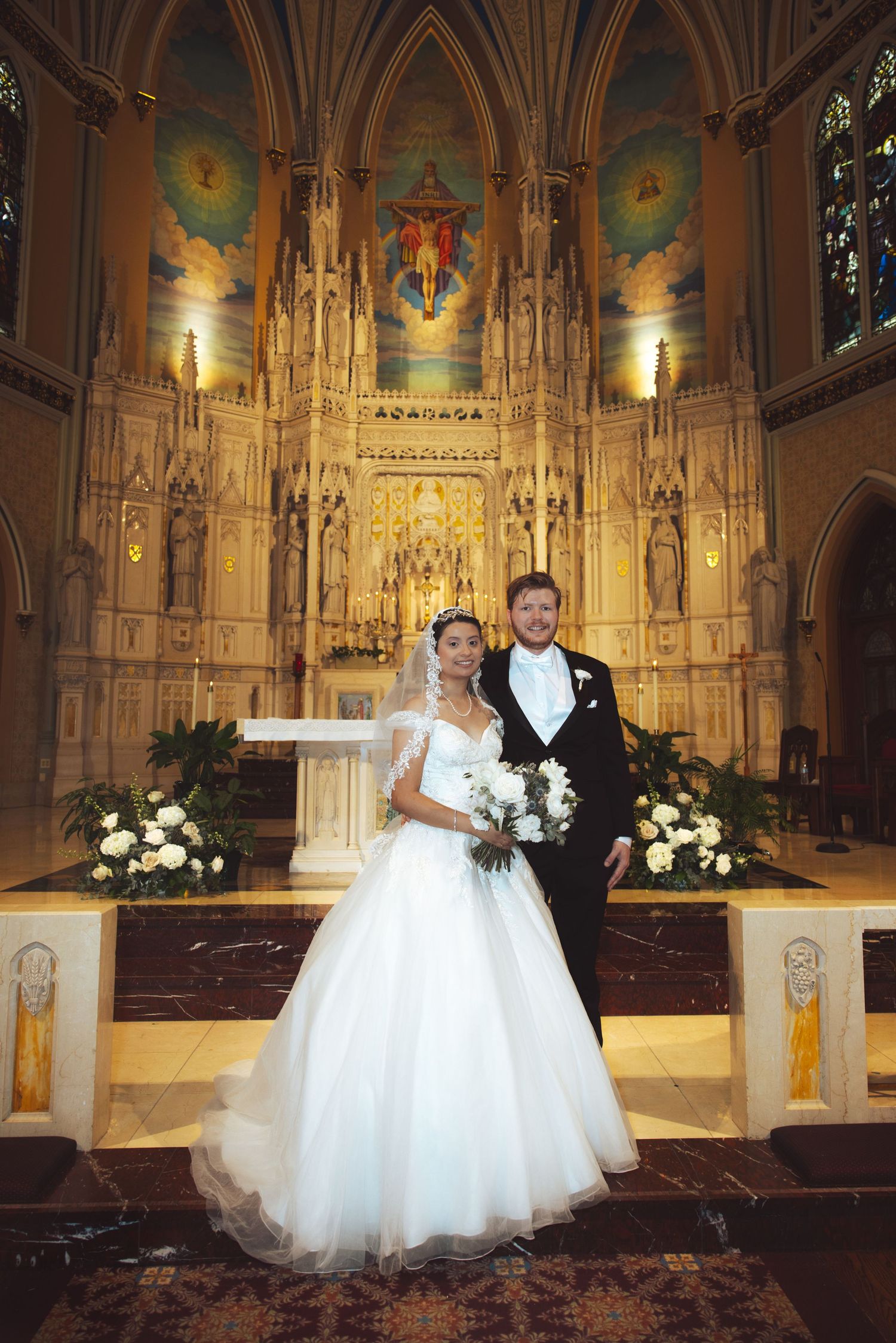 Bride and groom posing together at ornate church altar with Gothic architecture and floral arrangements.