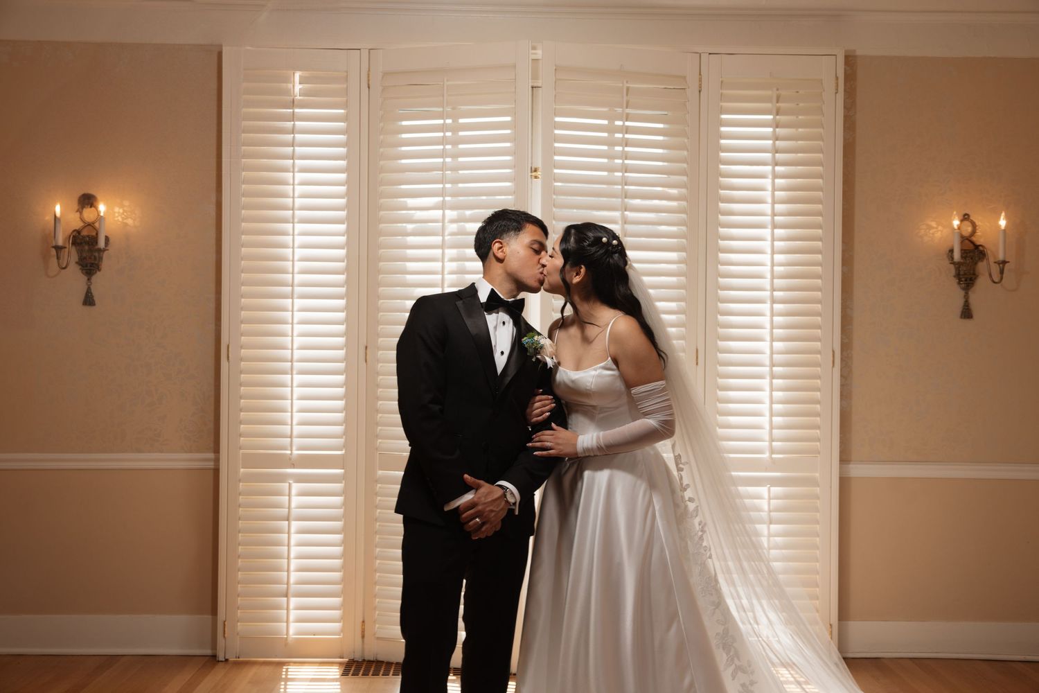 Bride and groom sharing a romantic kiss indoors in front of white shuttered doors on their wedding day.