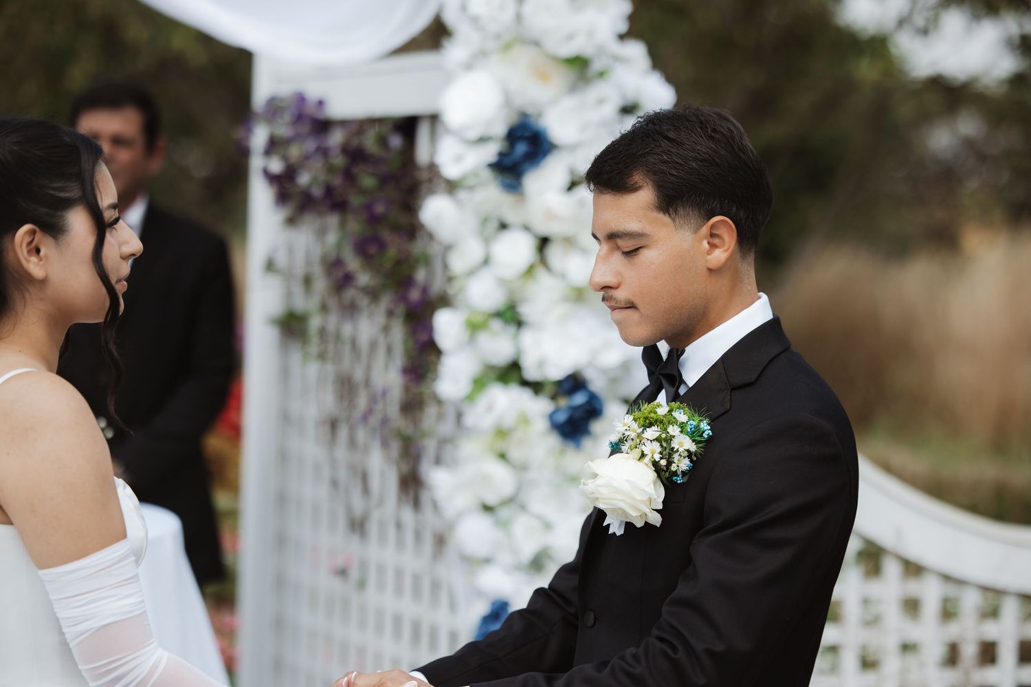 Groom in black tux holds bride's hands at outdoor wedding ceremony with floral arch.