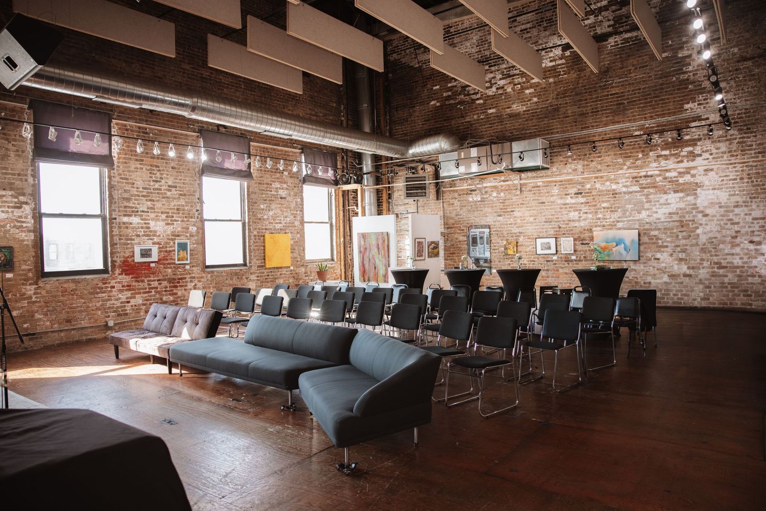 Spacious industrial loft venue with exposed brick walls, rows of black chairs, modern sofas, and hardwood floors.