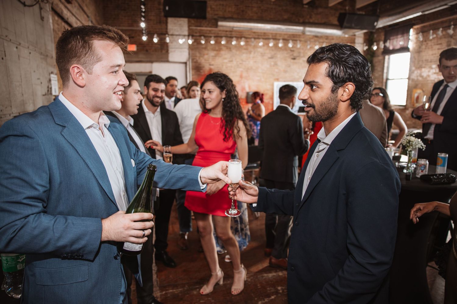Two men in suits shake hands at a social event while guests mingle in the background.
