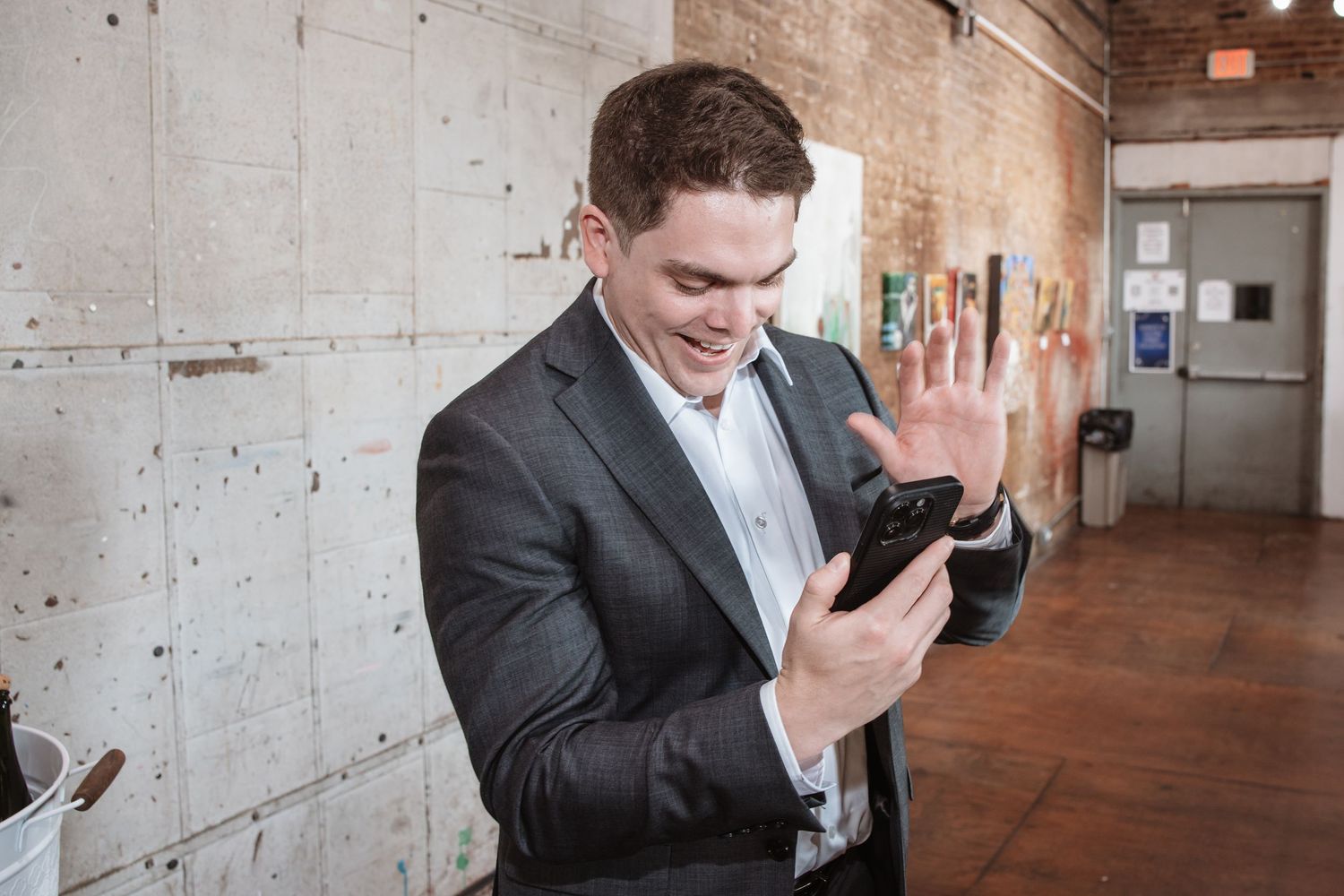 Smiling man in dark suit waves at his smartphone screen during a video call at an event.