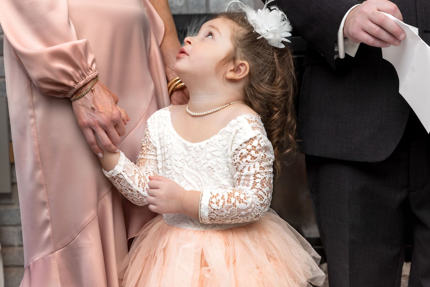 Adorable flower girl in white lace and blush tulle dress looking up at parents.