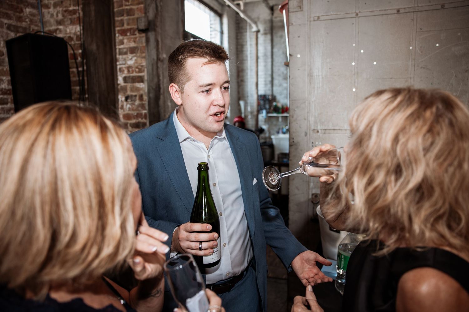 Man in blue suit holding champagne bottle conversing with guests at an indoor reception.