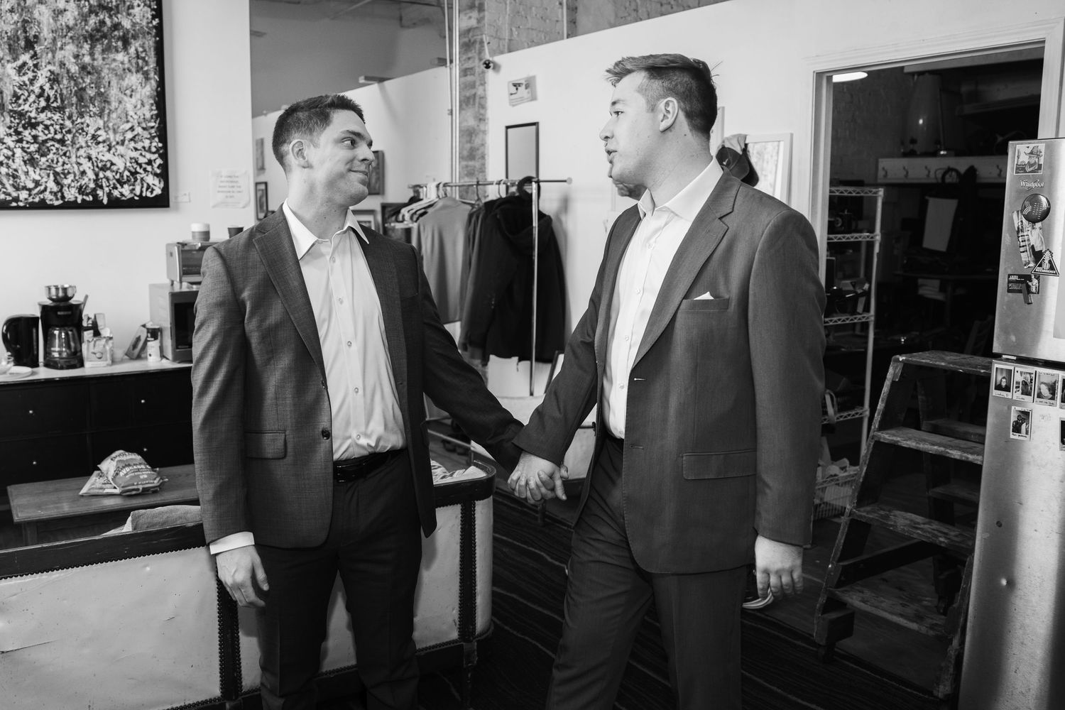 Two men in suits holding hands and smiling at each other in a black and white photo.