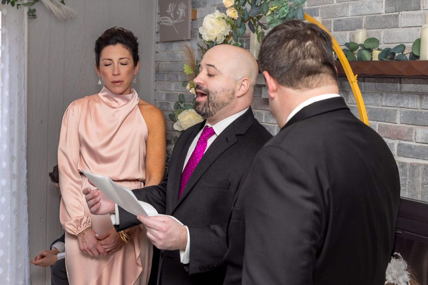 Woman in pink gown stands beside officiant reading vows at intimate indoor wedding ceremony.