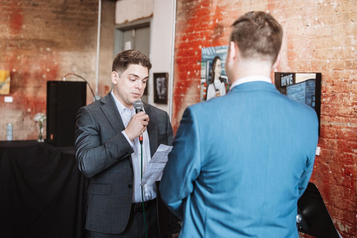 Man in dark suit speaks into microphone at elegant wedding reception with exposed brick walls.