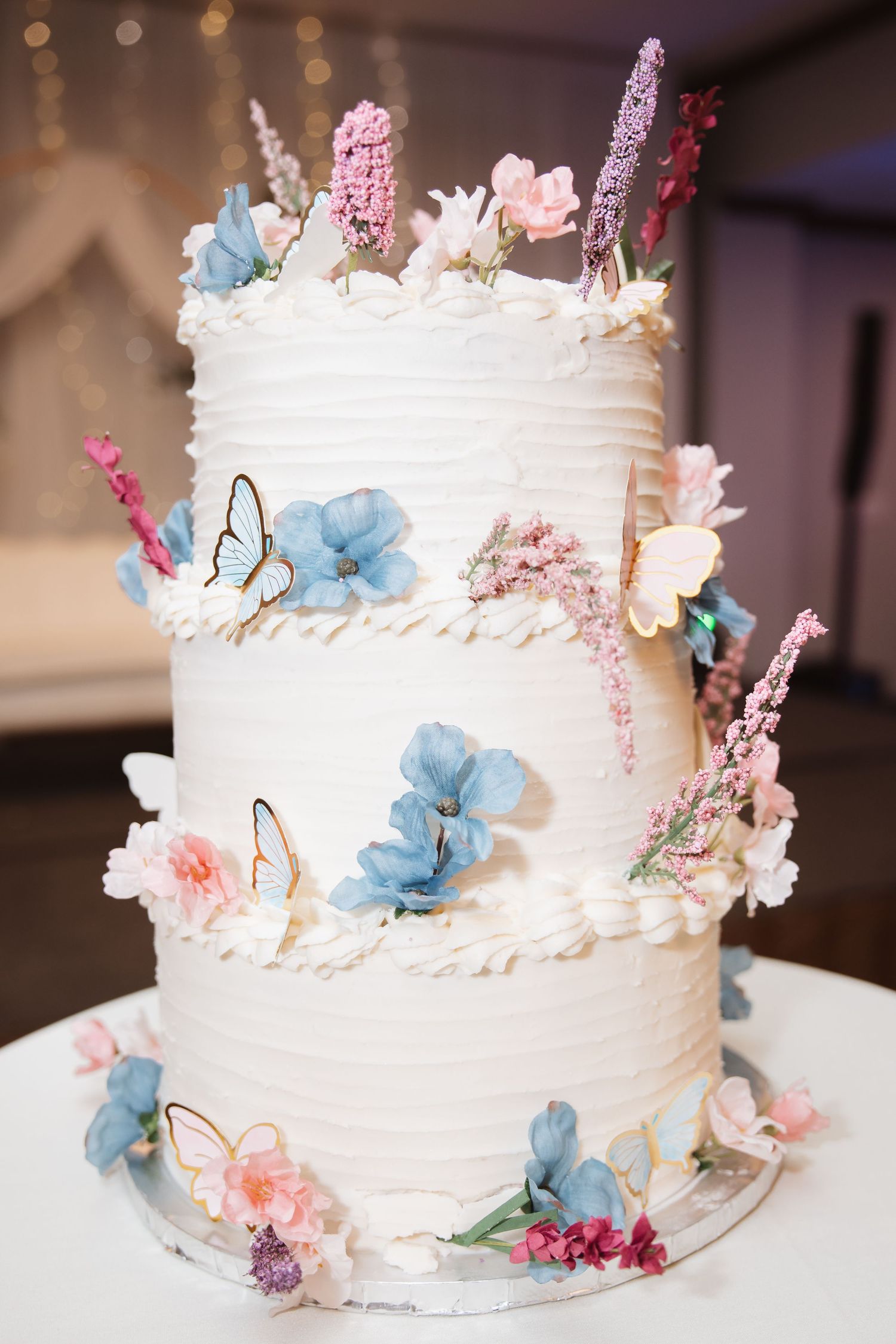 Elegant two-tier white wedding cake decorated with blue, pink flowers and butterfly accents.