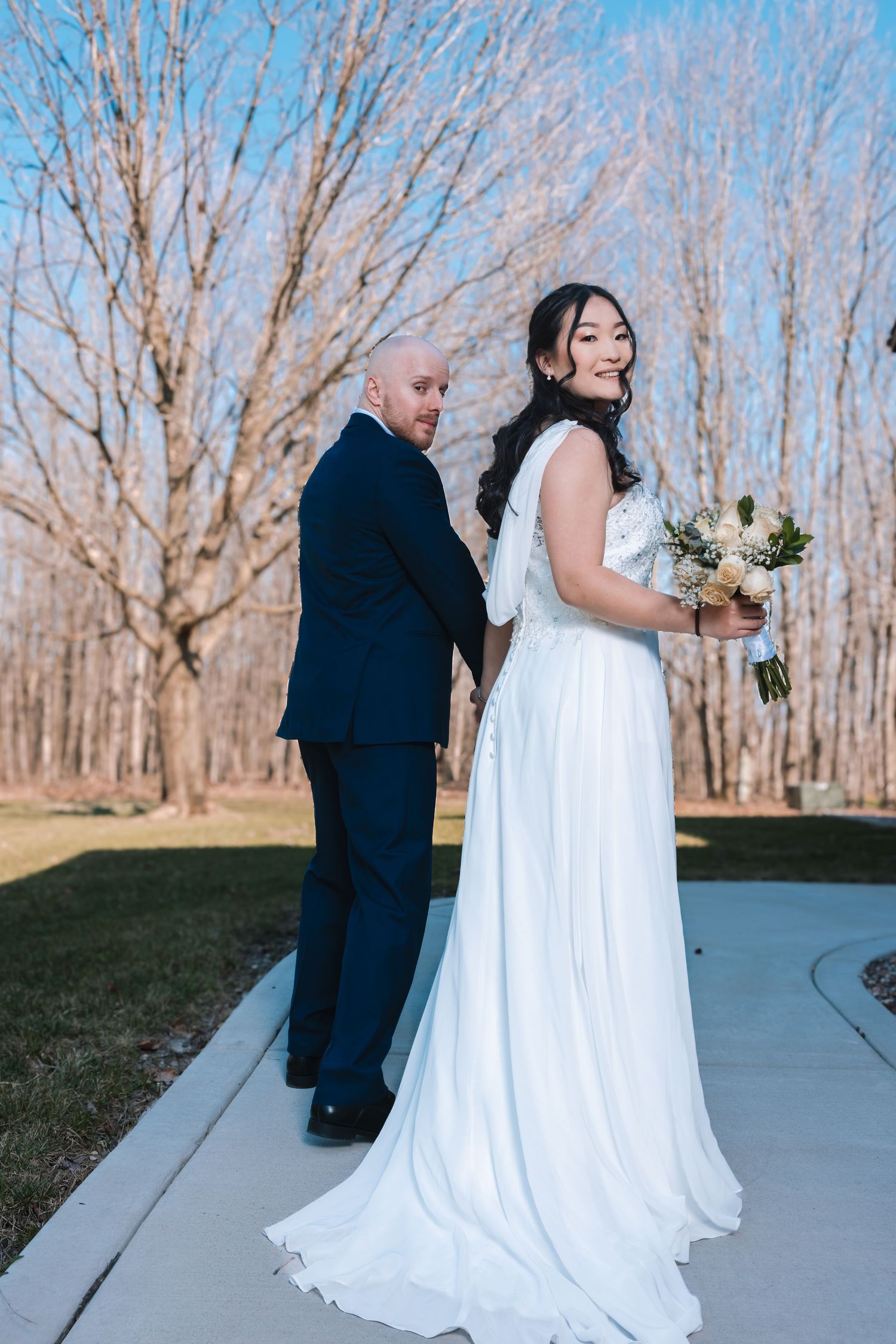 Bride in flowing white gown holds bouquet while groom in navy suit walks along outdoor path.
