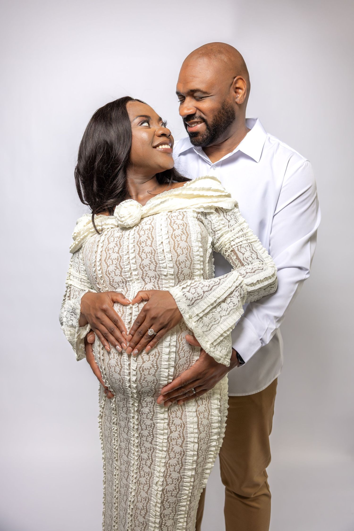 Smiling pregnant couple in studio, woman in white lace dress, man in white shirt, making heart on belly.