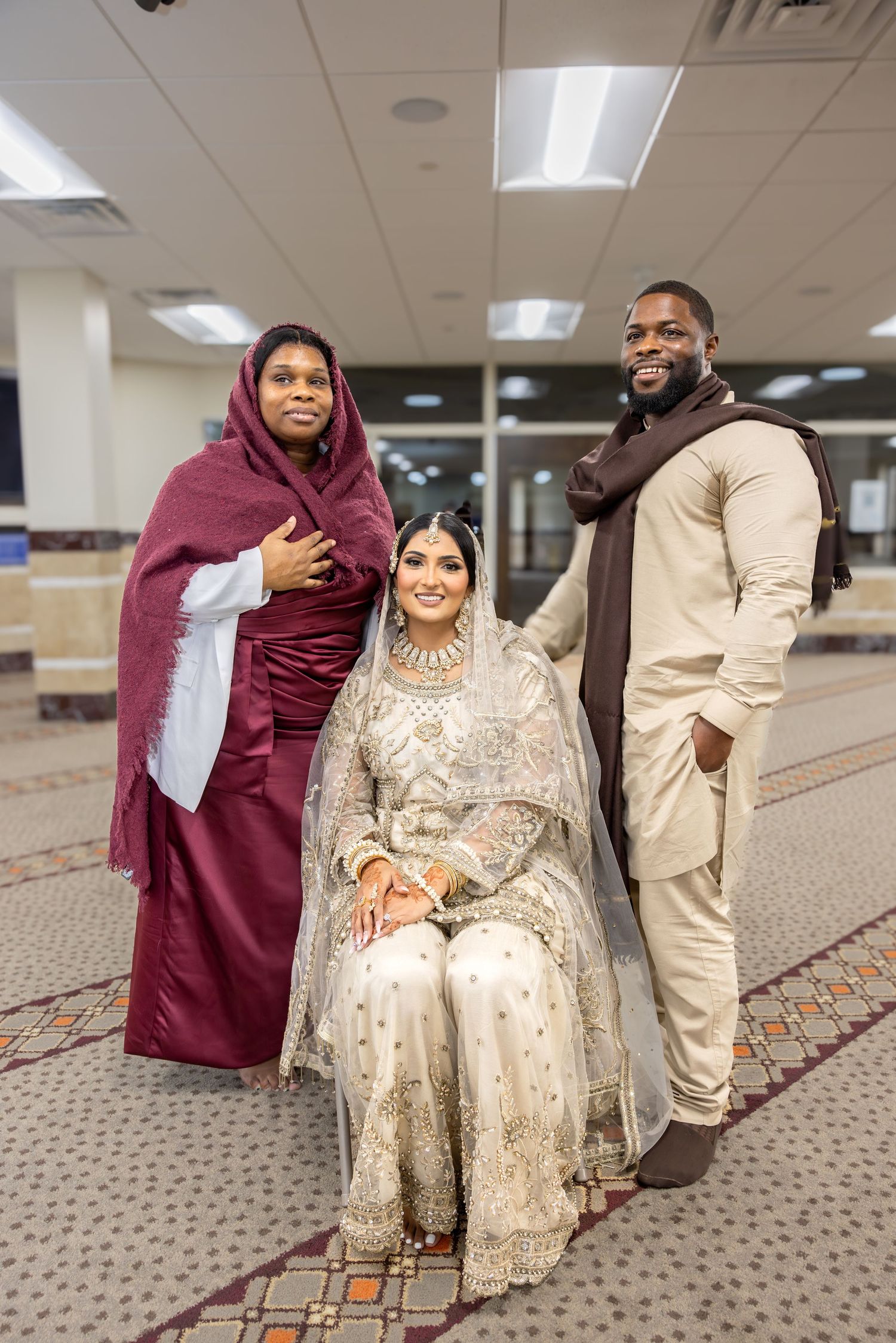 Muslim bride in white lehenga seated with groom and mother in maroon hijab inside a mosque.
